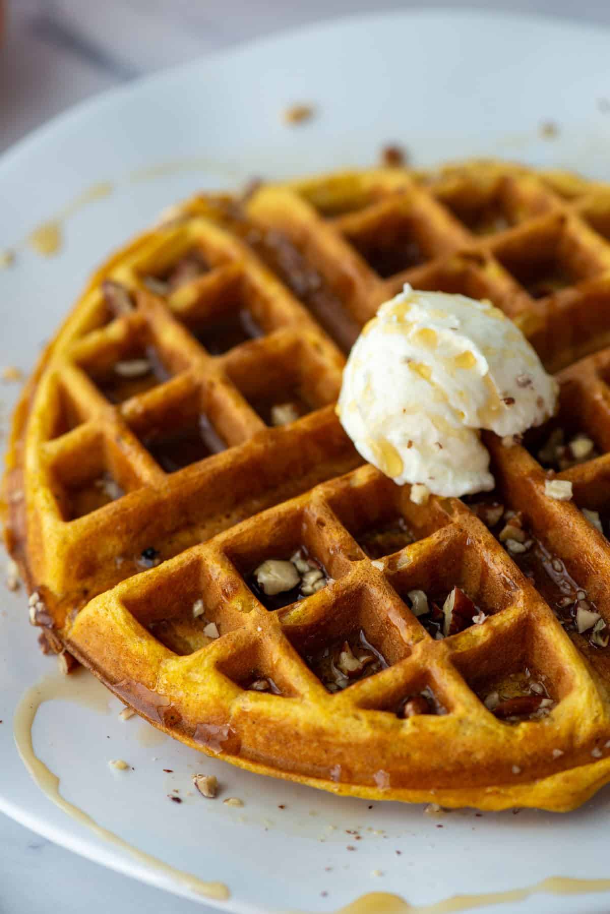 Overhead angled, partial view of a pumpkin spice waffle topped with maple syrup, chopped pecans and whipped butter on a plate.