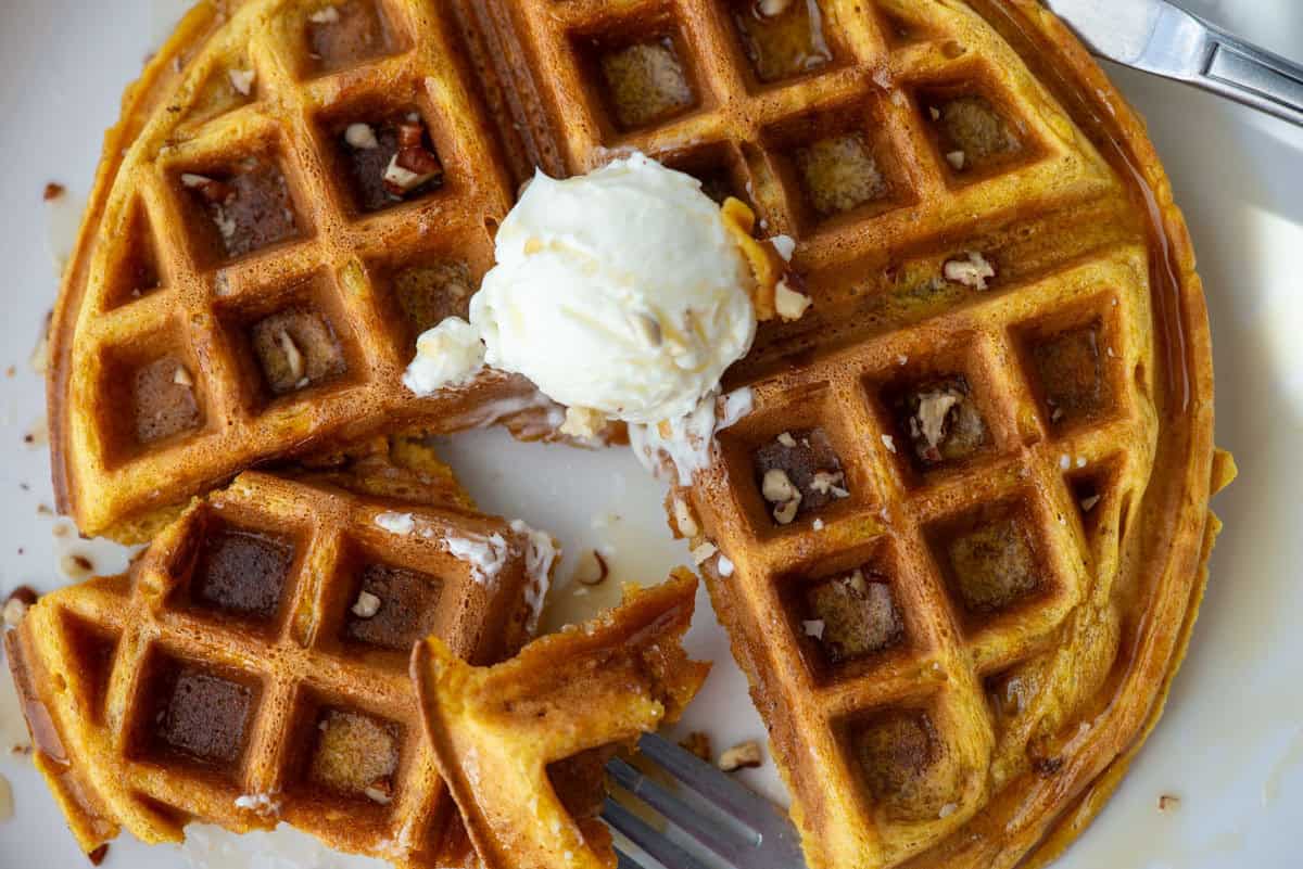Close up photo of a pumpkin spice waffle with a section cut out and a bite on a fork. You can see syrup and chopped pecans on top of the waffle and in the foreground as well as whipped butter on top of the waffle.