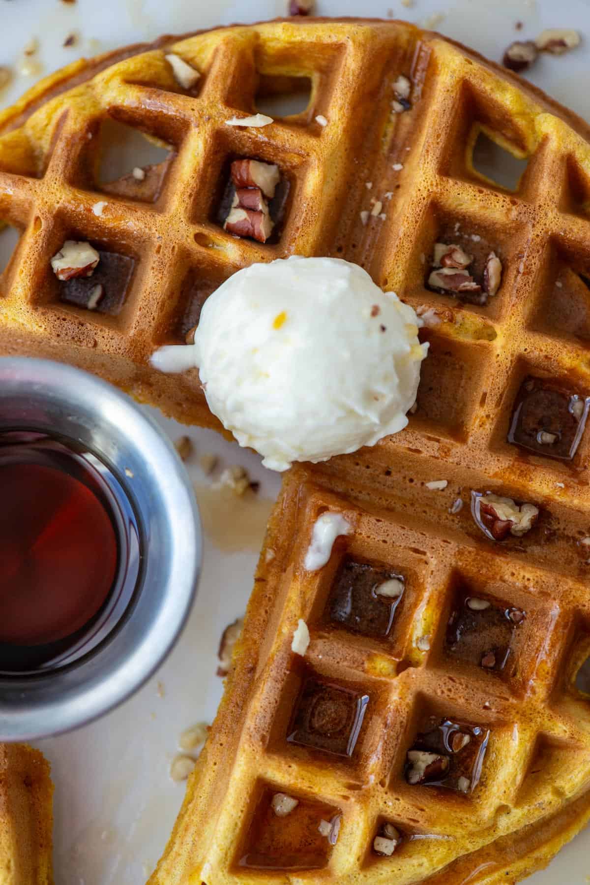 Overhead view of ¾ of a pumpkin spice waffle topped with whipped butter, maple syrup and chopped pecans. A small cup of syrup is also visible on the left.