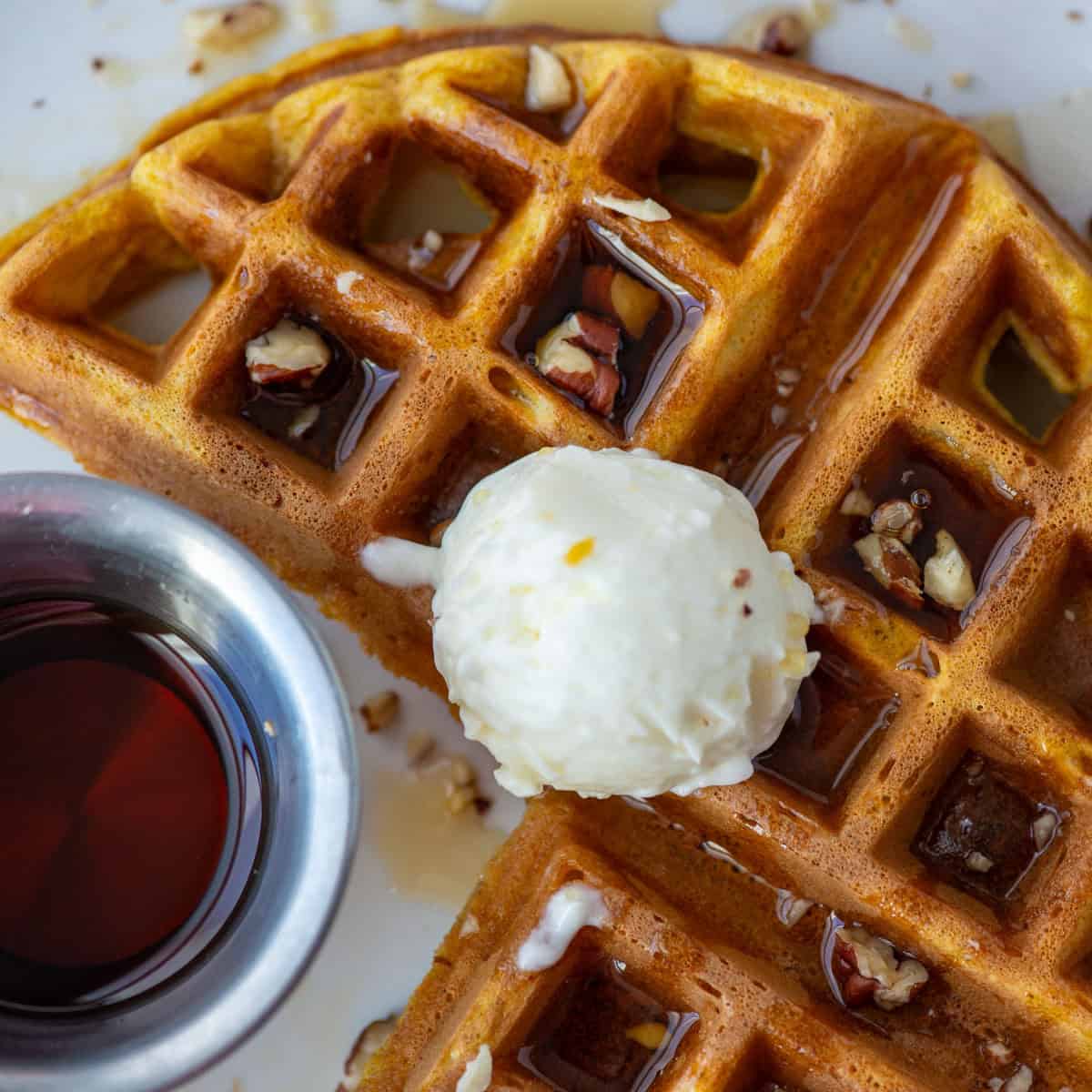 Overhead close up view of a partial pumpkin spice waffle topped with whipped butter, maple syrup and chopped pecans. A small cup of syrup is also visible on the left.