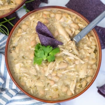 Overhead view of a bowl of turkey and white bean chili garnished with cilantro and a blue corn tortilla chip with a spoon sticking out of the side and tortilla chips scattered around the bowl