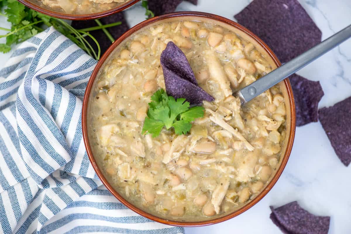 Overhead view of a bowl of turkey and white bean chili garnished with cilantro and a blue corn tortilla chip with a spoon sticking out of the side, tortilla chips scattered around the bowl and a napkin on the left side